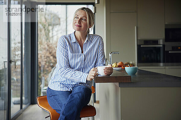 Nachdenkliche blonde Frau mit Kaffeetasse sitzt zu Hause in der Küche