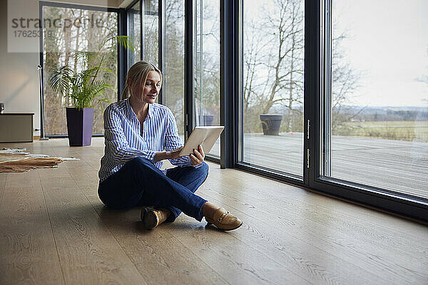 Frau sitzt mit Tablet-PC auf dem Boden vor einem Glasfenster zu Hause