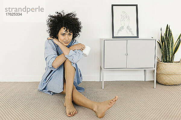 Lächelnde junge Frau mit Kaffeetasse sitzt zu Hause auf dem Boden neben einer weißen Wand