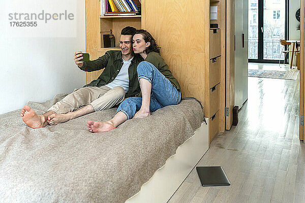 Glücklicher junger Mann  der im heimischen Schlafzimmer ein Selfie mit seiner Freundin per Smartphone macht