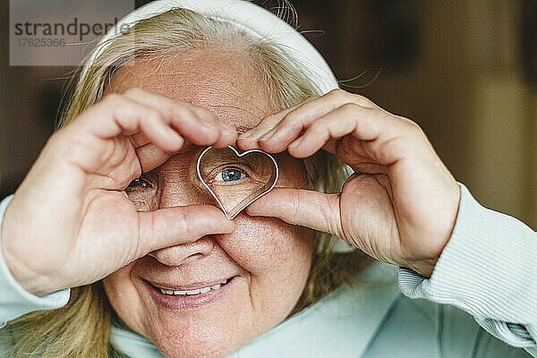 Lächelnde ältere Frau  die durch einen herzförmigen Keksausstecher schaut
