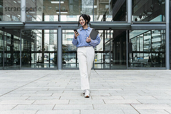 Lächelnde Geschäftsfrau mit Laptop und Mobiltelefon vor dem Bürogebäude