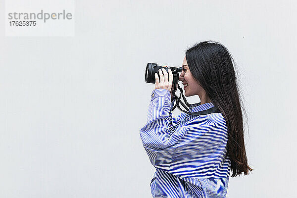Happy woman photographing through camera standing in front of white wall