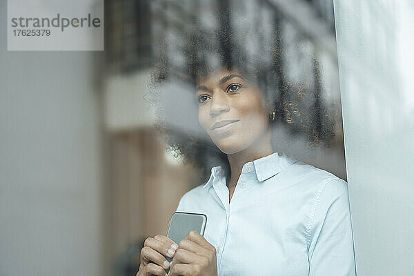 Lächelnde Frau mit Smartphone durch Glasfenster gesehen