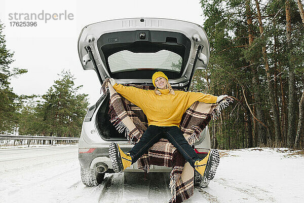 Glückliche Frau in warmer Kleidung sitzt im Winter im Kofferraum eines Autos