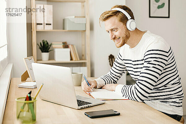 Lächelnder Mann mit kabellosen Kopfhörern  der im Wohnzimmer auf seinen Laptop schaut