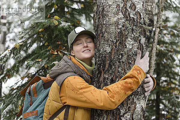 Junge Frau mit geschlossenen Augen umarmt Baum im Winterwald