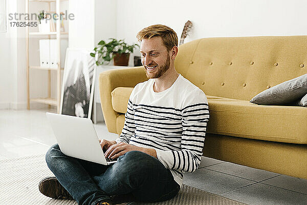 Lächelnder junger Mann mit Laptop  der zu Hause mit gekreuzten Beinen vor dem Sofa sitzt