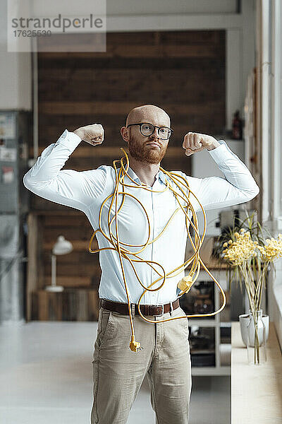Selbstbewusster junger Geschäftsmann  bedeckt mit Kabel  der im Büro Muskeln spielen lässt