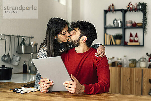 Mann hält Tablet-PC und küsst Freundin zu Hause in der Küche
