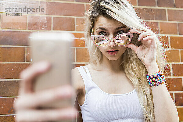 Blond teenage girl wearing sunglasses taking selfie through smart phone in front of brick wall