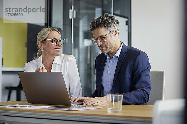 Lächelnde Geschäftsfrau blickt auf Kollegen  der im Büro am Laptop arbeitet