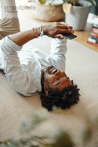 Smiling man using mobile phone lying on floor in living room