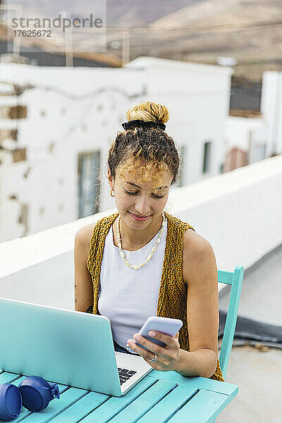 Lächelnde Frau  die über ihr Mobiltelefon im Internet surft und mit einem Laptop am Tisch auf dem Dach sitzt