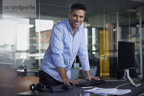 Lächelnder Geschäftsmann lehnt am Schreibtisch am Arbeitsplatz