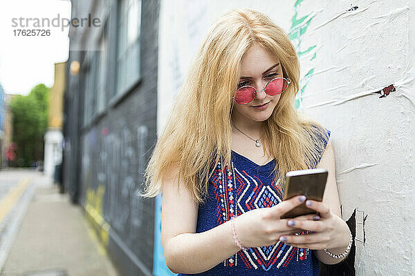 Blond teenage girl wearing sunglasses surfing net through mobile phone by wall