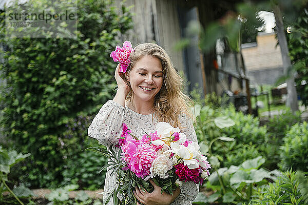 Schöne lächelnde blonde Frau mit rosa Blume steht im Garten