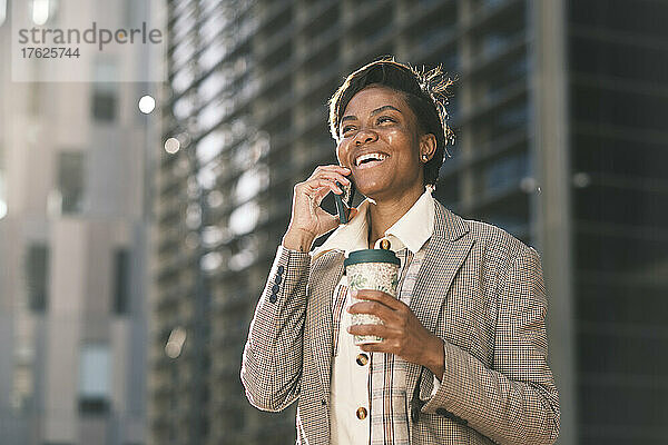 Fröhliche Geschäftsfrau hält Kaffeetasse in der Hand und telefoniert an einem sonnigen Tag
