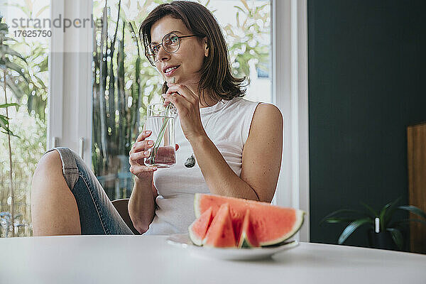 Nachdenkliche Frau trinkt zu Hause Wasser mit Wassermelone auf dem Tisch