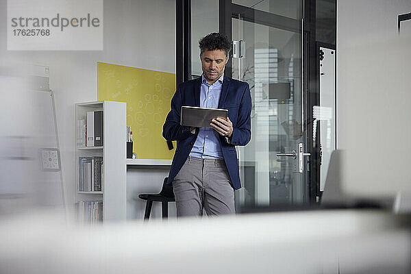Geschäftsmann stützt sich am Arbeitsplatz auf einen Tablet-PC und lehnt an einer Glastür