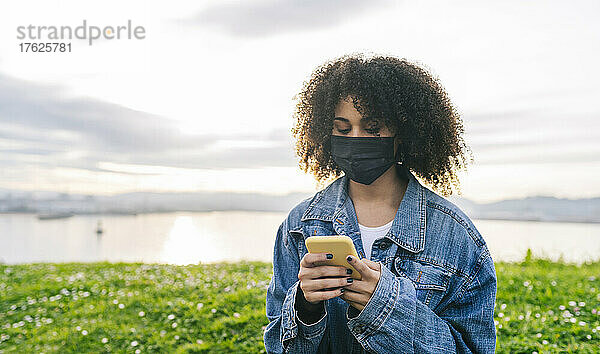 Frau mit schützender Gesichtsmaske benutzt Mobiltelefon in der Natur