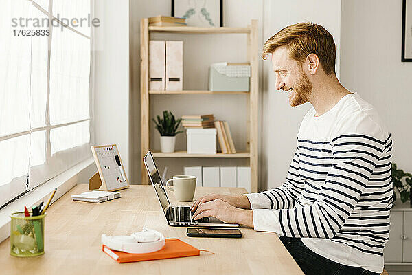Lächelnder junger Mann  der am Tisch im Wohnzimmer sitzt und auf dem Laptop tippt