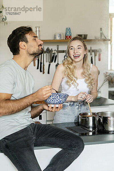 Fröhliche junge Frau bereitet Essen zu und schaut ihrem Freund beim Essen mit geschlossenen Augen zu  wie er zu Hause auf der Kücheninsel sitzt