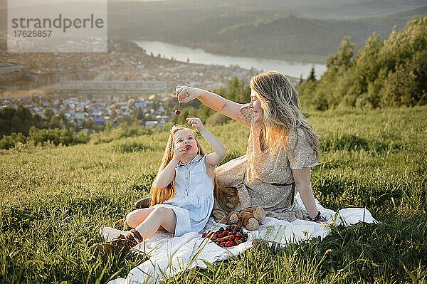 Glückliche Mutter füttert Tochter  die auf Gras sitzt  Beeren