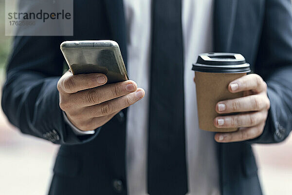 Hände eines Geschäftsmannes  der sein Smartphone benutzt und dabei eine Einwegkaffeetasse in der Hand hält