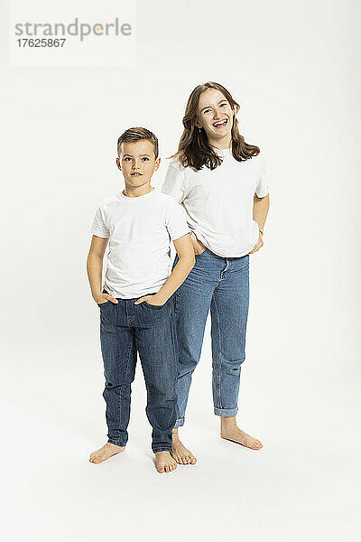 Happy girl and brother standing with hands in pockets against white background