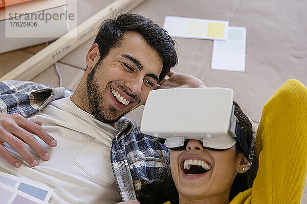 Glückliche Frau mit VR-Brille liegt neben ihrem Freund bei Renovierungsarbeiten zu Hause