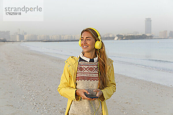Glückliche Frau mit Smartphone und gelben Kopfhörern am Strand
