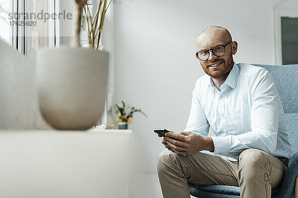 Smiling businessman with mobile phone sitting on chair in office