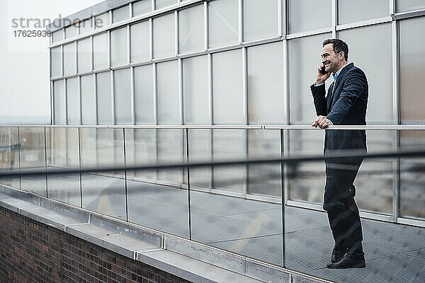 Smiling businessman talking on smart phone by glass railing on balcony