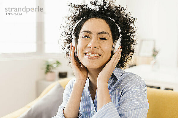 Glückliche Frau mit lockigem Haar  die zu Hause Musik über kabellose Kopfhörer hört