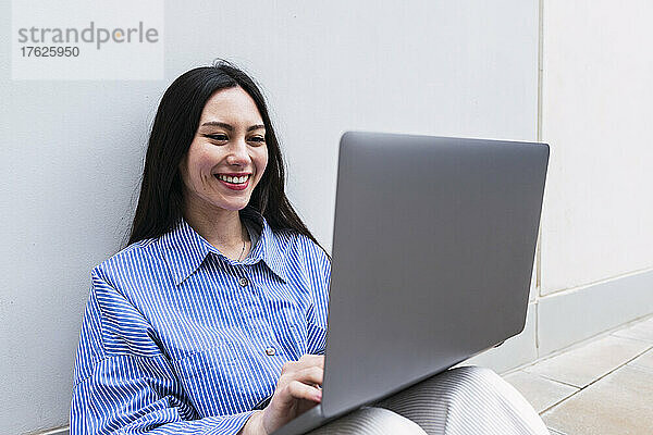 Lächelnde Geschäftsfrau mit Laptop sitzt vor weißer Wand