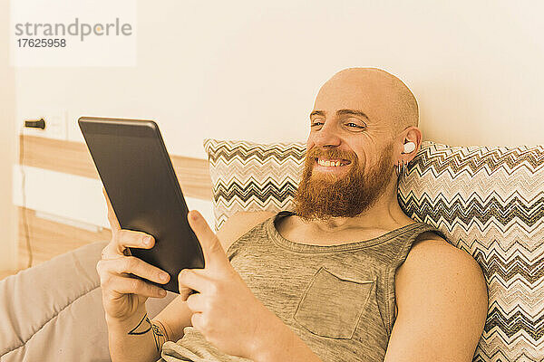 Smiling man using tablet PC and listening music through in-ear headphones lying on bed at home