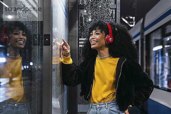 Fröhliche junge Frau mit kabellosen Kopfhörern sucht nachts an der Straßenbahnhaltestelle auf der Karte