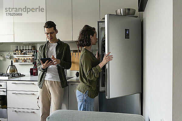 Junger Mann benutzt Tablet-PC  während Frau zu Hause den Kühlschrank in der Küche öffnet