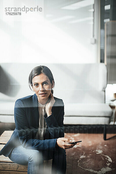 Geschäftsfrau mit Mobiltelefon sitzt auf einer Holzkiste im Büro