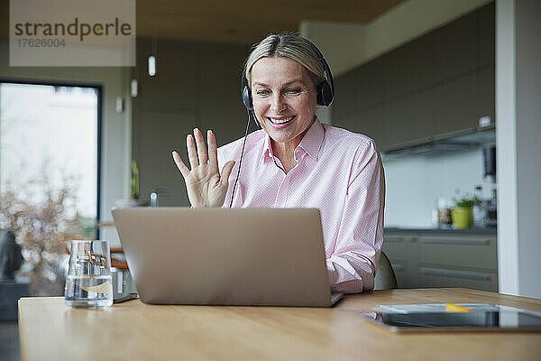 Glückliche Frau mit Kopfhörern winkt bei Videoanruf über Laptop am Tisch mit der Hand
