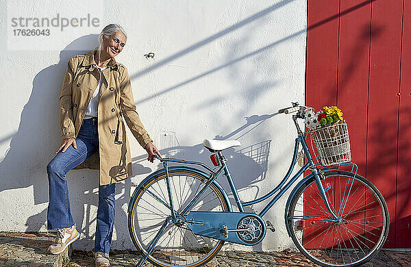 Lächelnde Frau mit Fahrrad steht an sonnigem Tag vor der Wand