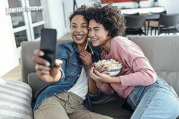 Fröhliche Freunde machen Selfies mit dem Handy und sitzen auf dem Sofa im Wohnzimmer