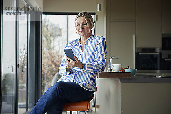 Blonde Frau surft mit Smartphone im Internet und sitzt in der Küche