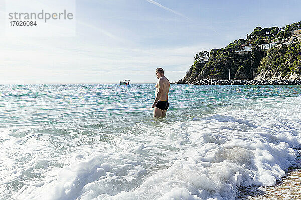 Mann mit nacktem Oberkörper steht am Strand im Meer