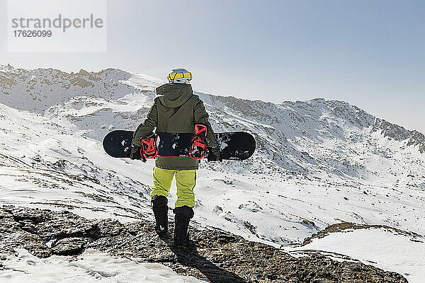 Junger Mann steht mit Snowboard auf einem Felsen