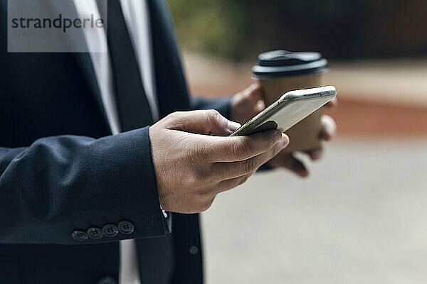 Hände eines Geschäftsmannes  der sein Smartphone benutzt und dabei eine Einwegkaffeetasse in der Hand hält