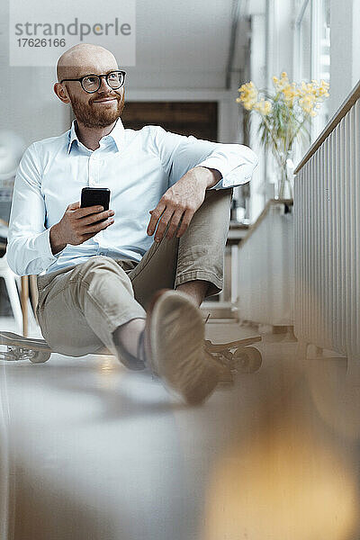 Businessman with mobile phone sitting on skateboard in office