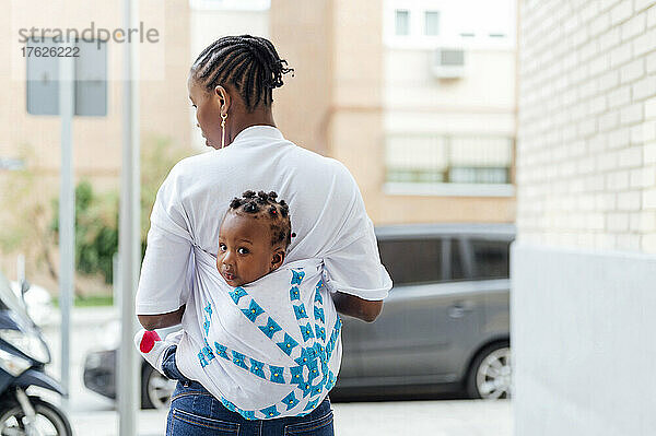 Junge Frau trägt süße Tochter in Kanga-Babytrage und geht an Mauer entlang