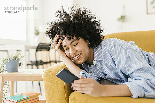 Glückliche Frau mit der Hand im Haar  die zu Hause auf dem Sofa ihr Mobiltelefon benutzt